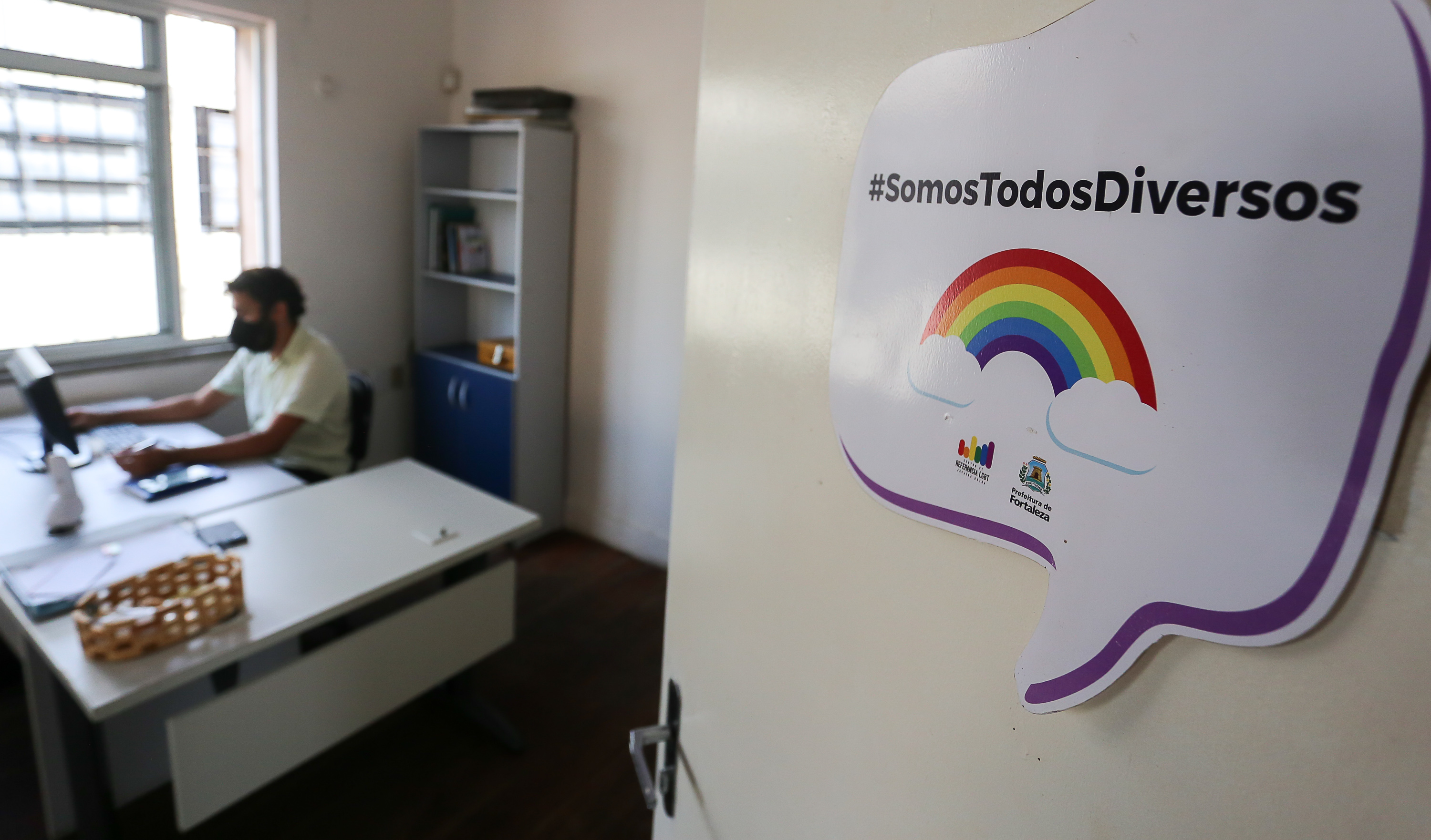 a foto mostra a porta de uma sala dentro do Centro de Referência LGBT Janaína Dutra com um homem trabalhando em uma mesa